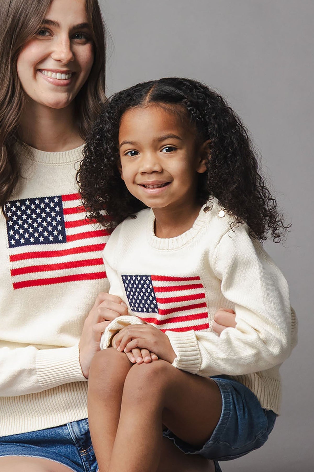 A woman and a young girl wear matching ivory sweaters featuring an American flag design. The sweaters are made from soft organic cotton, perfect for family outings and celebrations.
