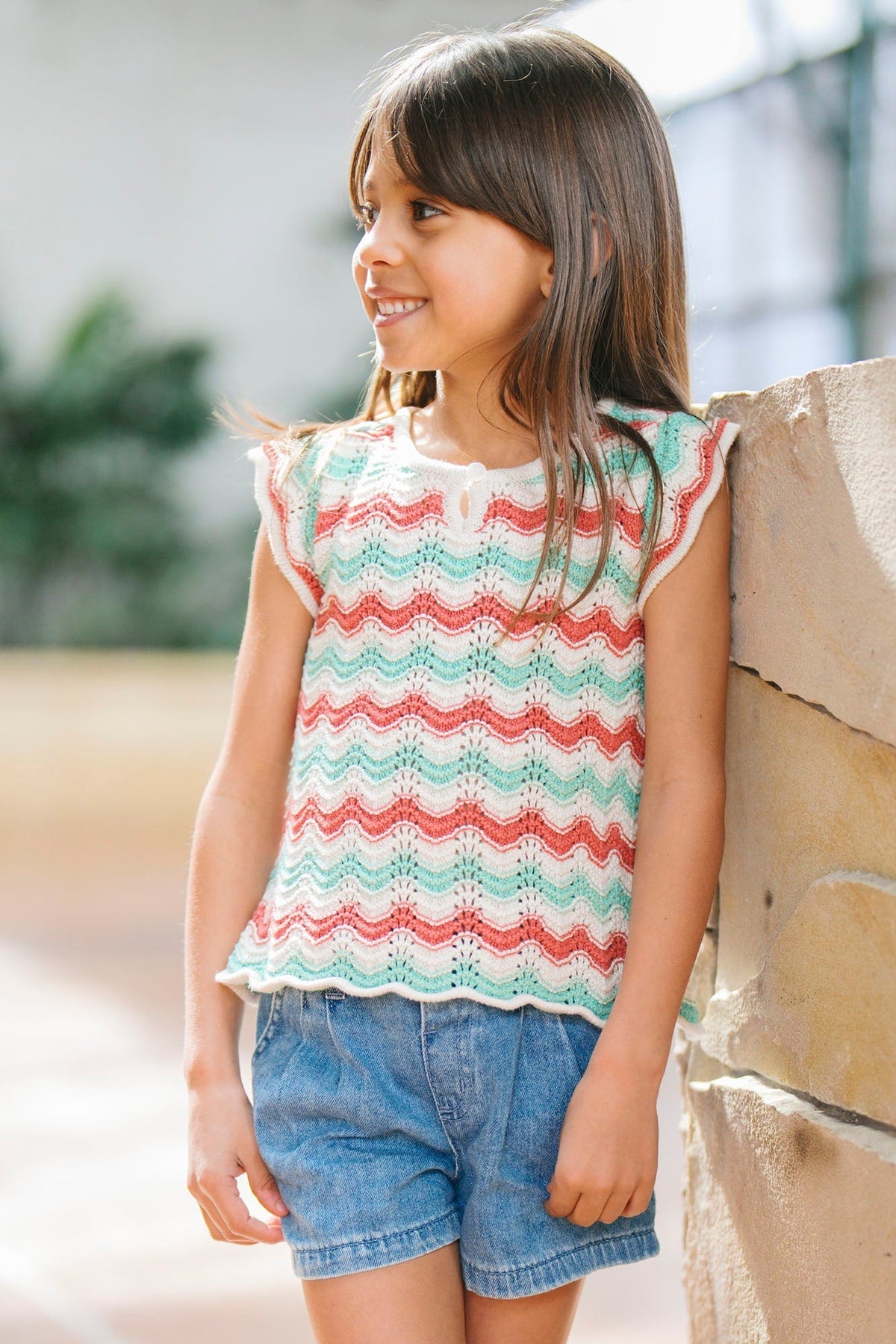 A young girl smiles while wearing a colorful, striped organic cotton sweater top with flutter sleeves, paired with denim shorts, set against a bright, airy indoor background.