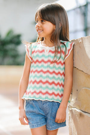 A young girl smiles while wearing a colorful, striped organic cotton sweater top with flutter sleeves, paired with denim shorts, set against a bright, airy indoor background.