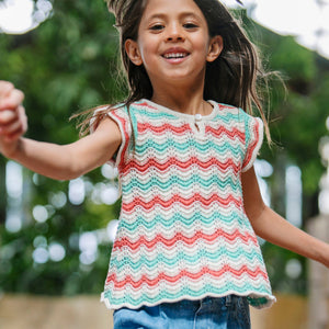 A young girl joyfully models a colorful, organic cotton sweater top featuring wavy stripes in red, teal, and cream. The airy design includes flutter sleeves, perfect for warm weather.