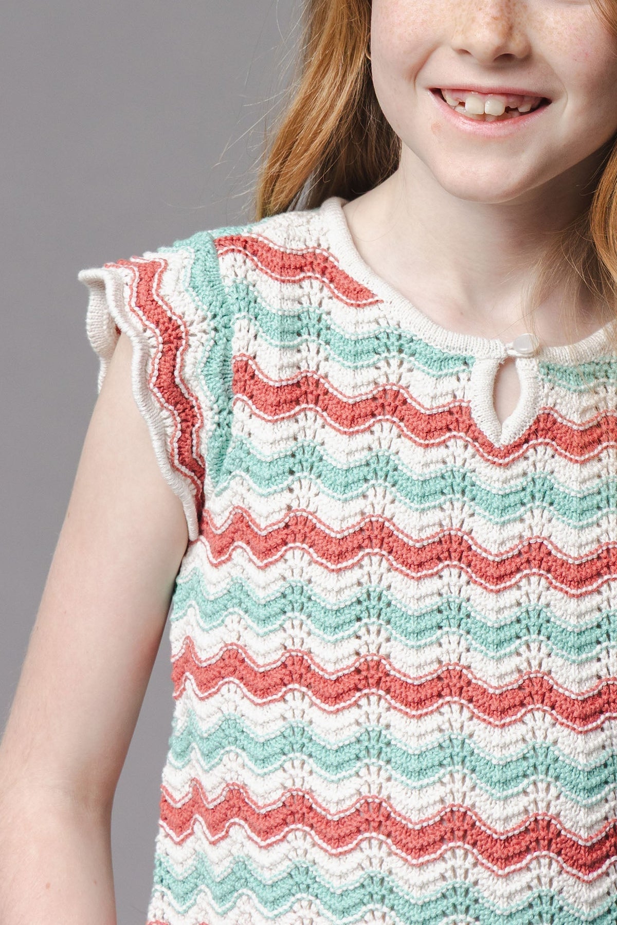A girl wears a colorful, textured knit sweater top with wavy stripes in mint green, coral, and cream, featuring flutter sleeves and a delicate keyhole closure. Ideal for warm weather.