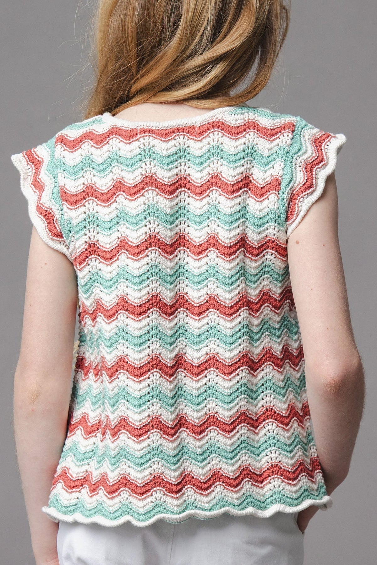 A child models a lightweight organic cotton sweater top featuring a wavy, multicolored pattern in red, green, and white, with flutter sleeves and a comfortable fit for warm weather.