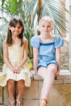 Two young girls sit on a stone ledge surrounded by tropical plants. One wears a yellow striped dress, while the other sports a blue puff sleeve romper.