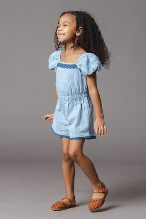 A young girl wears a blue organic cotton seersucker romper featuring puff sleeves and crochet trim. She smiles joyfully, showcasing the rompers comfortable fit and playful design.