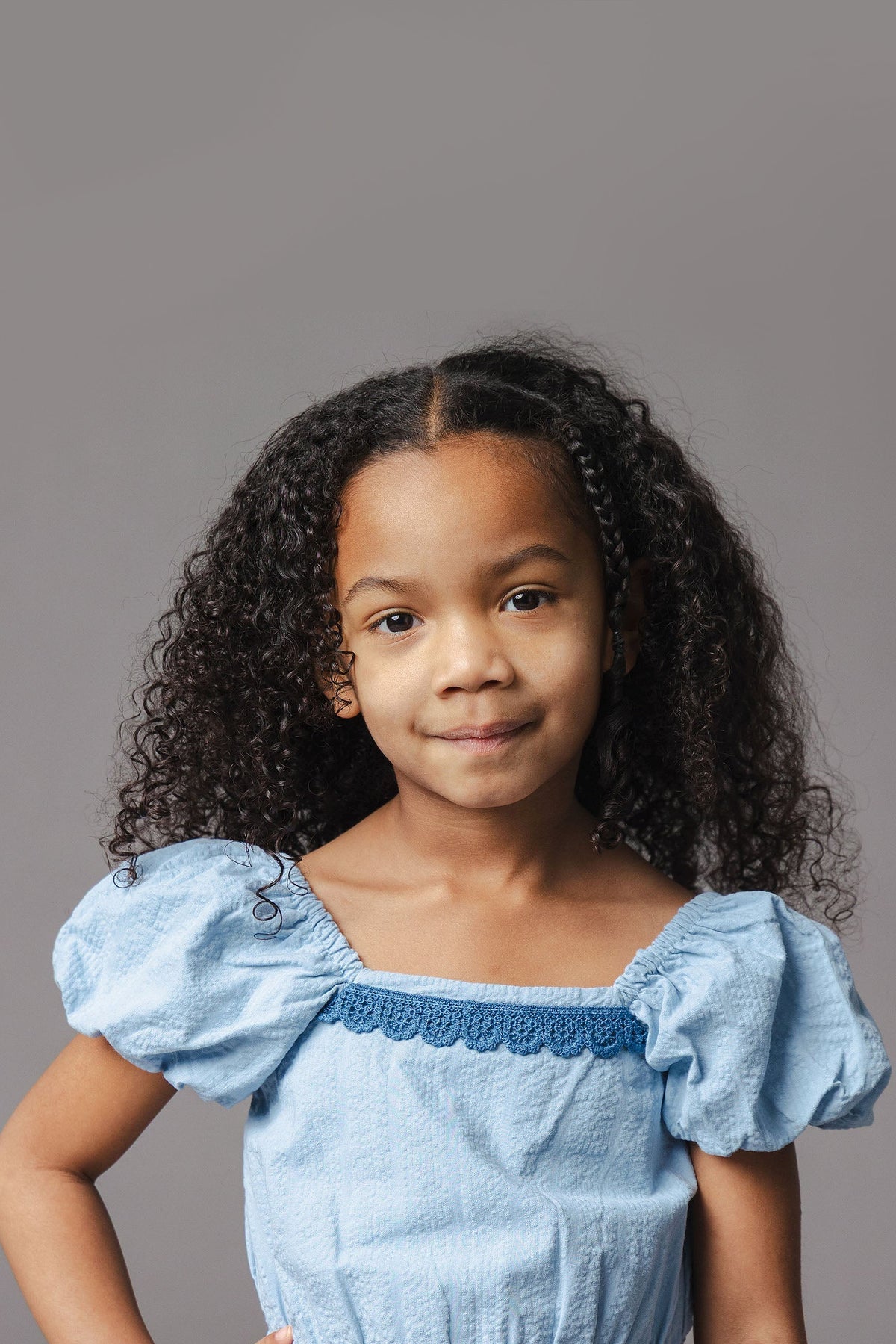 A young girl with curly hair wears a light blue organic cotton romper featuring puff sleeves and a crochet trim. The background is a neutral gray, highlighting her outfit.