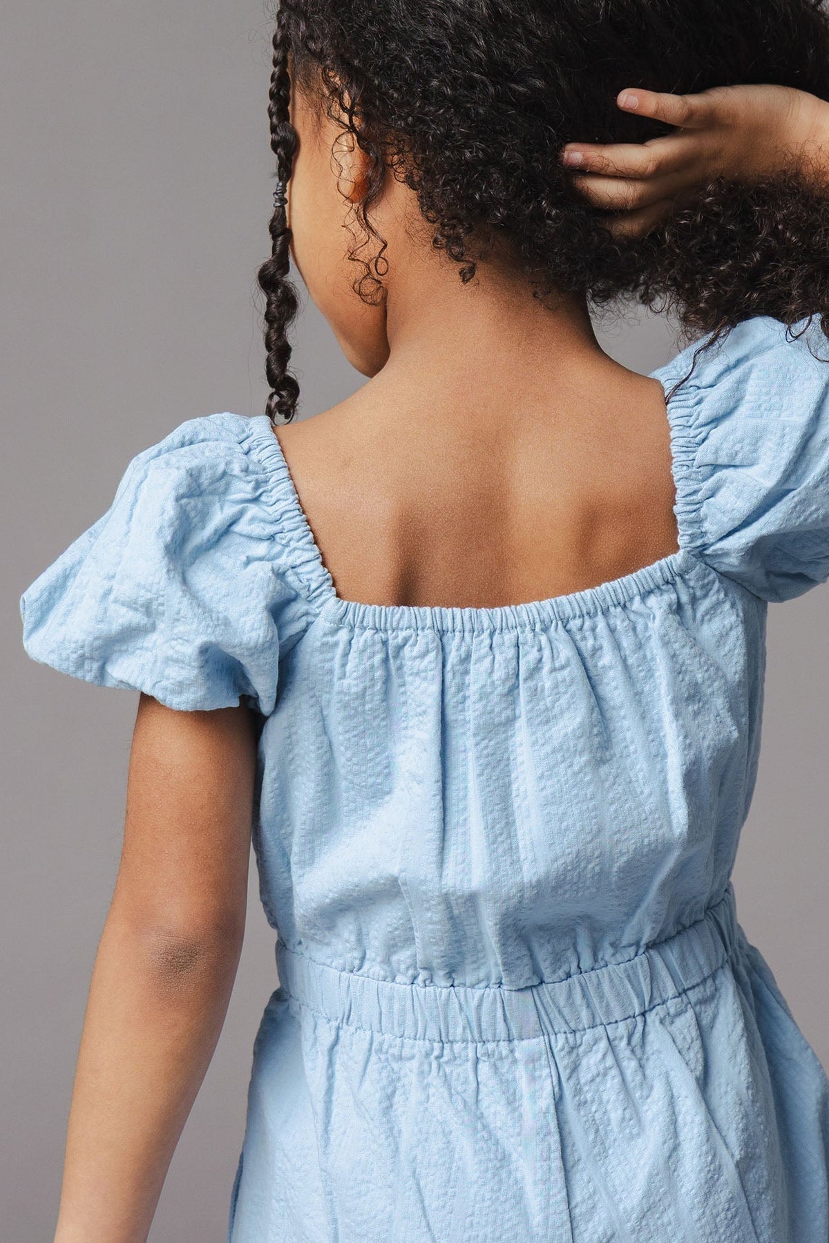 A young child is seen from behind, wearing a light blue organic cotton romper with puff sleeves. The romper features an elastic waistband, showcasing a comfortable and stylish fit.