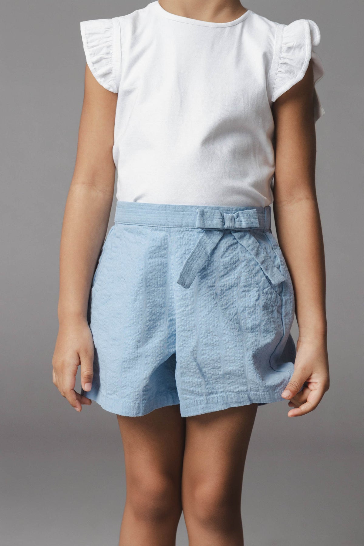 A child wears a white short-sleeve shirt and light blue organic cotton seersucker shorts featuring a faux belt and decorative bow. The outfit is stylish and perfect for warm weather.
