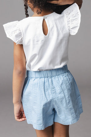 A young girl stands with her back to the camera, wearing a white blouse with eyelet sleeves and light blue organic cotton seersucker shorts, showcasing a comfortable and stylish outfit.