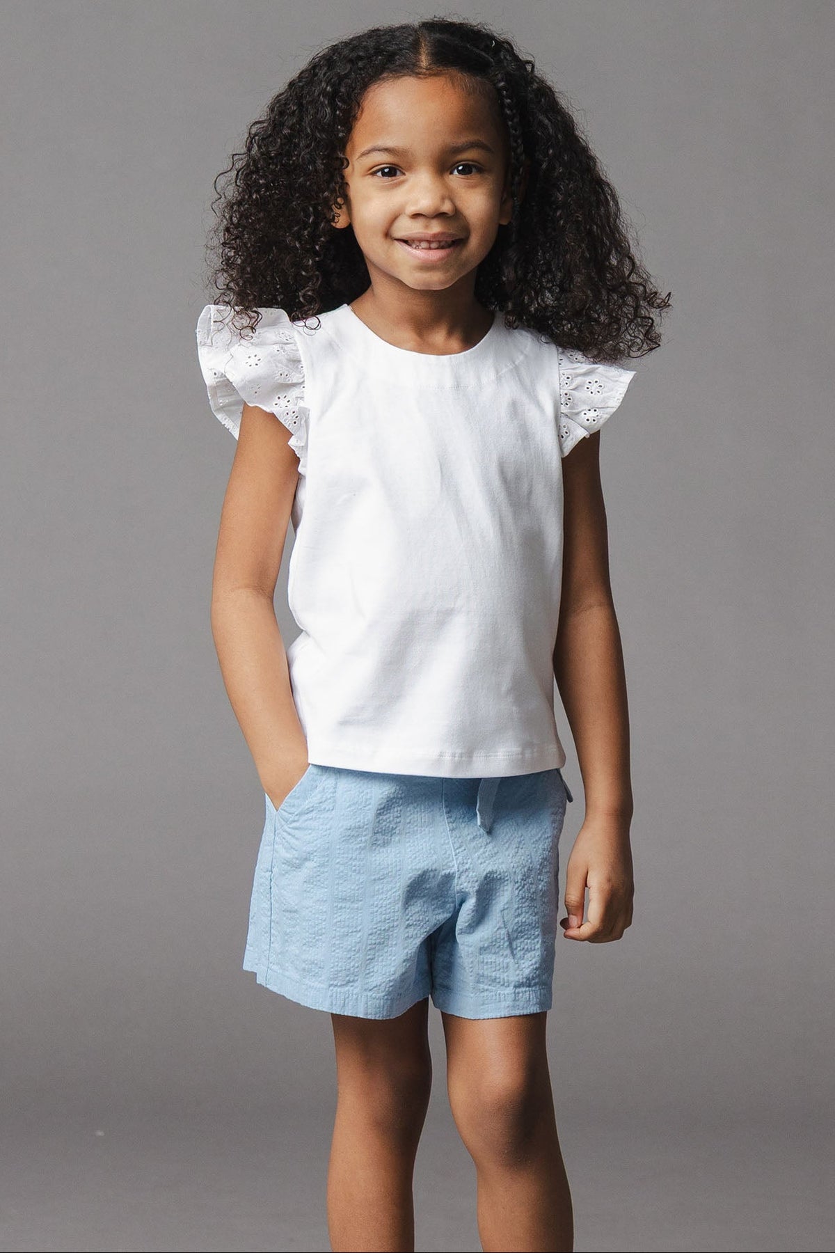 A young girl stands smiling, wearing a white sleeveless top with ruffled sleeves and blue organic cotton seersucker pull-on shorts. The background is a neutral gray, highlighting her outfit.