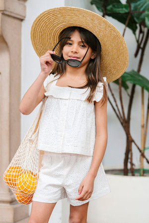 A young girl wears a natural eyelet tank top and matching shorts, paired with a wide-brimmed straw hat and sunglasses, standing in a sunny outdoor setting with greenery.