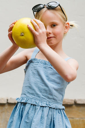 A young girl holds a large yellow fruit while wearing a light blue, organic cotton seersucker dress with ruffle edges. She accessorizes with stylish sunglasses, set against a neutral background.