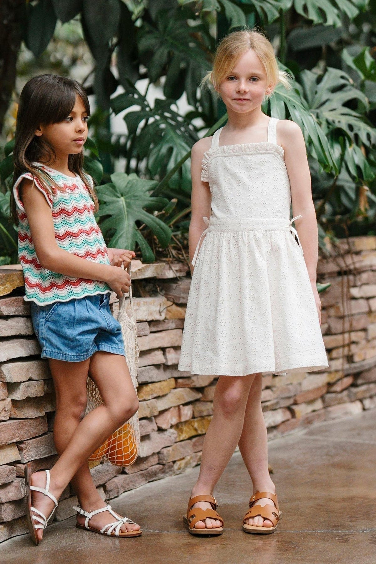 Two young girls stand outdoors near a stone wall, one wearing a colorful zigzag tank top and denim shorts, while the other models a white organic cotton eyelet dress with.