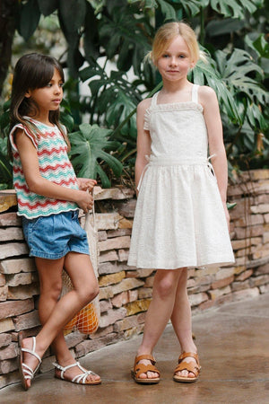 Two young girls stand outdoors near a stone wall, one wearing a colorful zigzag tank top and denim shorts, while the other models a white organic cotton eyelet dress with.