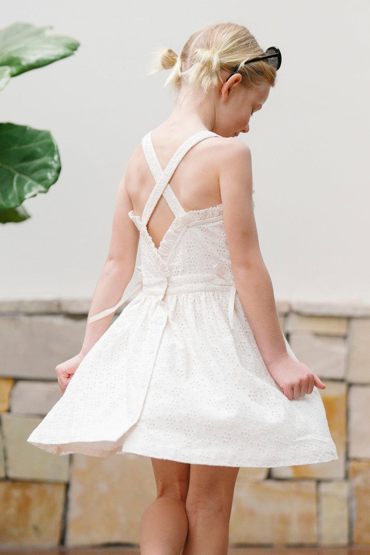 A young girl models a light beige organic cotton dress with ruffle edges and cross back straps, standing in a bright, outdoor setting near a leafy plant.