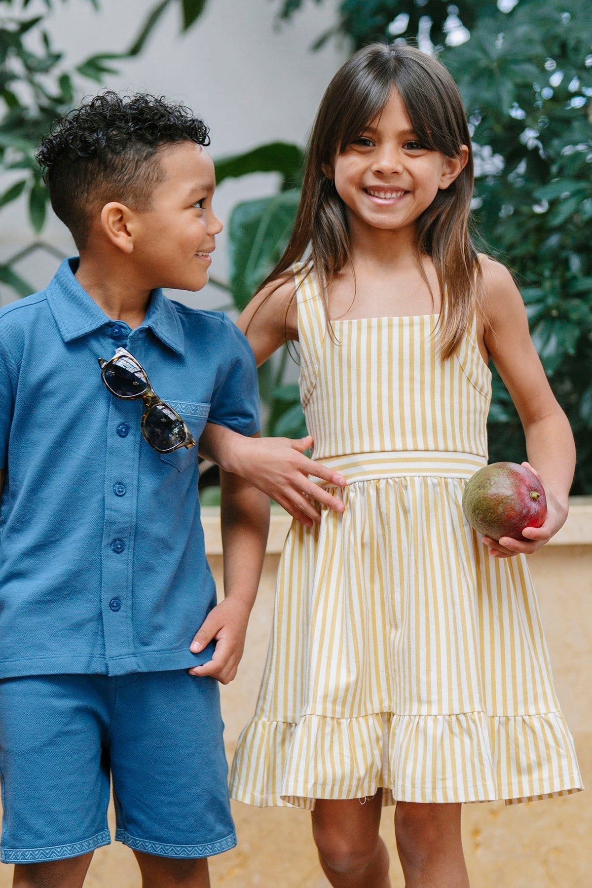 A young boy in a blue button-up shirt and shorts stands next to a girl wearing a yellow striped sundress with a flounce hem, both smiling in a lush outdoor.