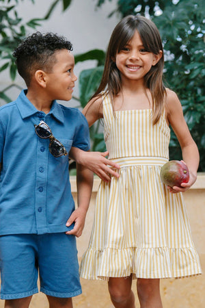 A young boy in a blue button-up shirt and shorts stands next to a girl wearing a yellow striped sundress with a flounce hem, both smiling in a lush outdoor.