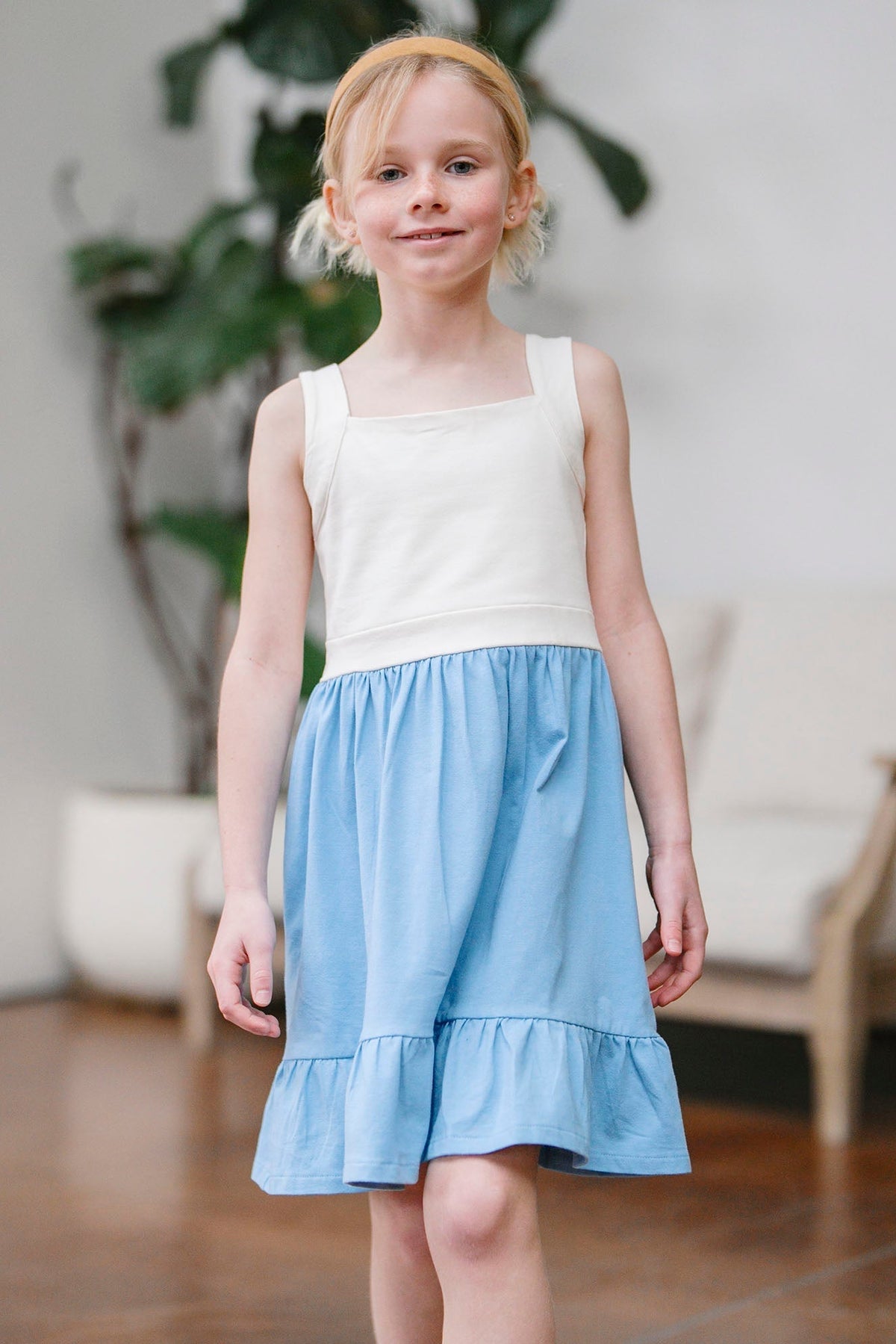 A young girl walks confidently in a blue and cream organic cotton sundress, featuring a flounce hem and a shirred waist. The setting is a bright, airy room with plants.