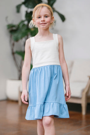 A young girl walks confidently in a blue and cream organic cotton sundress, featuring a flounce hem and a shirred waist. The setting is a bright, airy room with plants.