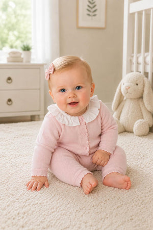 A smiling baby sits on a soft, neutral rug wearing a pale pink organic cotton romper with a delicate ruffle collar.