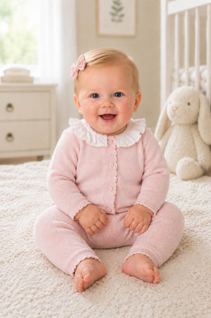 A smiling baby sits on a soft, textured rug, wearing a pale pink organic cotton romper with a delicate ruffle collar. The cozy nursery setting features a plush bunny.