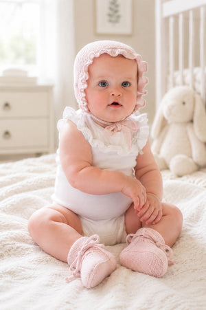 A baby sits on a cozy white blanket, wearing a pale pink bonnet with ruffled edges and matching knitted booties.