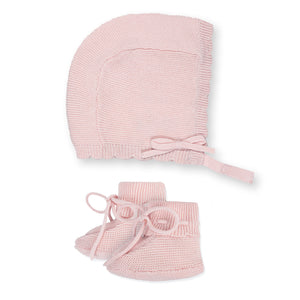 A pale pink knitted bonnet with a tie closure is displayed alongside matching booties featuring a round shape and bow detail, both made from soft, organic cotton for baby comfort.