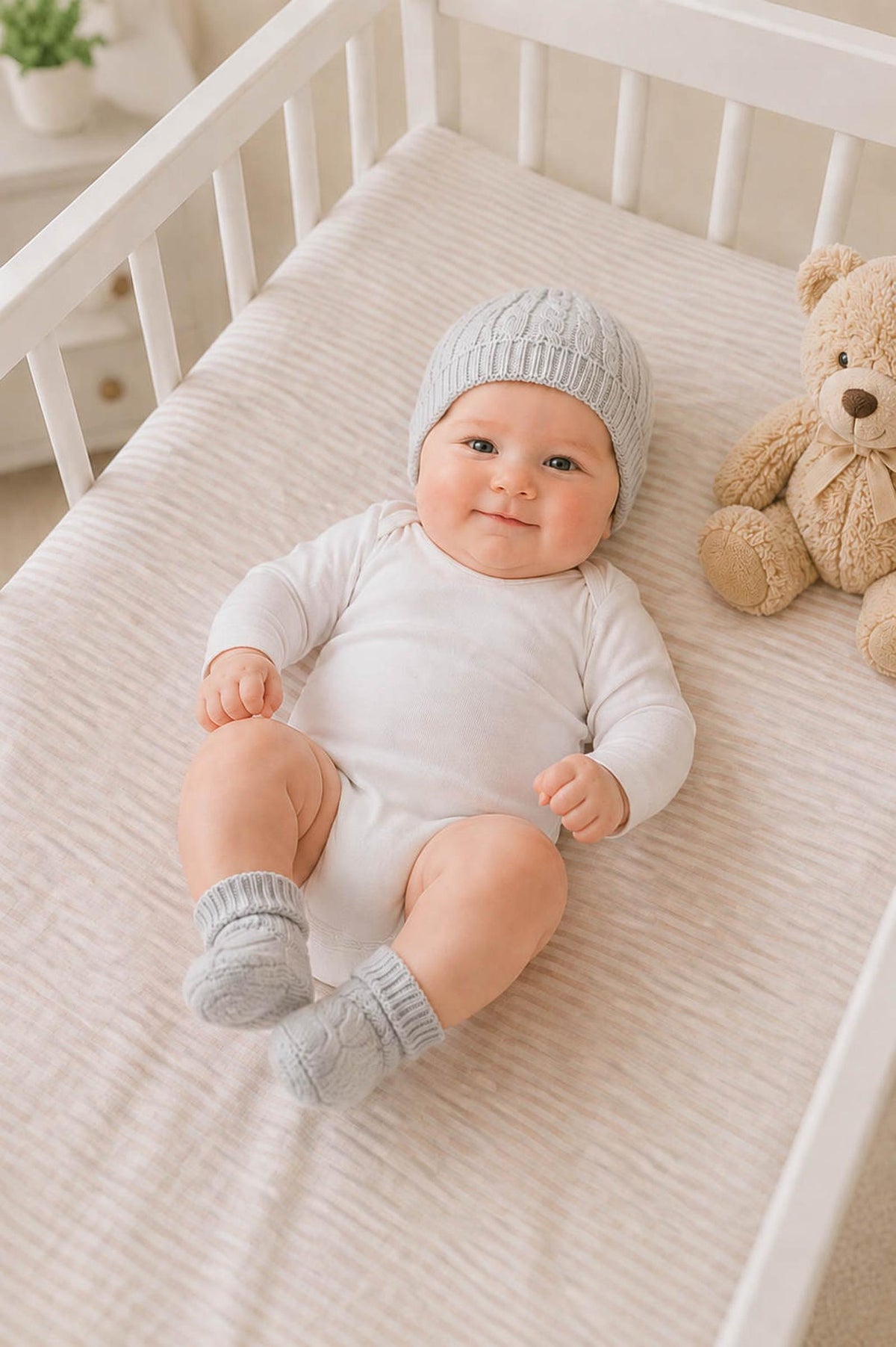 A cute baby wearing a light gray knitted beanie and matching booties lies on a soft, striped blanket in a crib, with a teddy bear nearby.