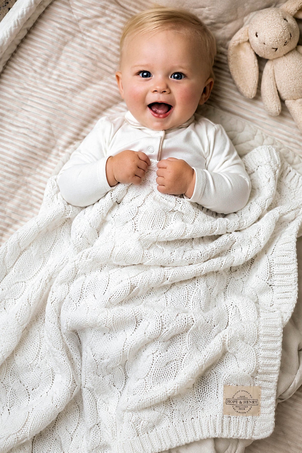 A joyful baby lies on a soft, textured blanket, wrapped in a cozy white cable knit organic cotton blanket. A plush bunny toy rests nearby, enhancing the serene atmosphere.