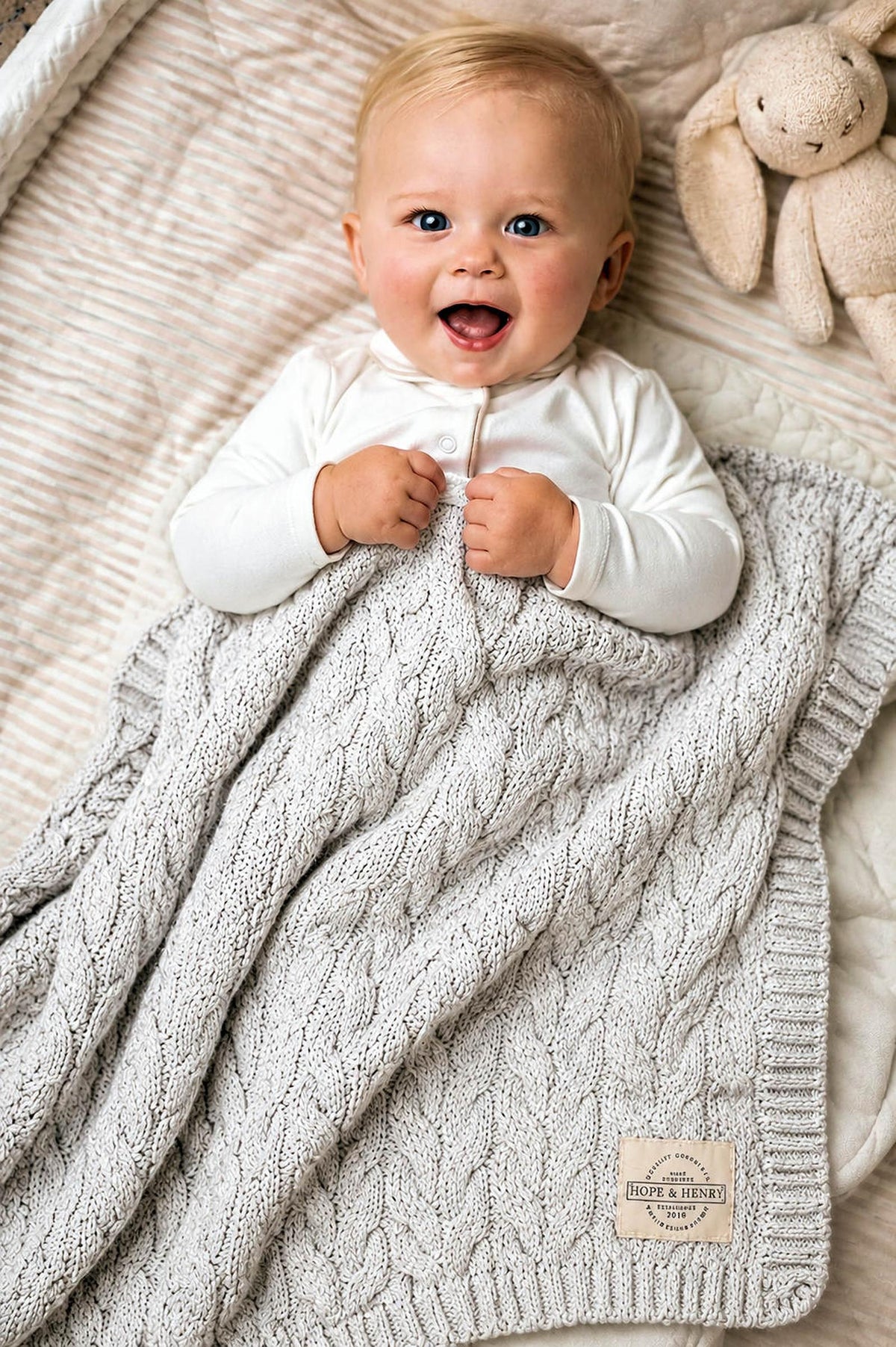A happy baby lies on a soft, textured blanket, wrapped in a light gray organic cotton cable knit blanket.