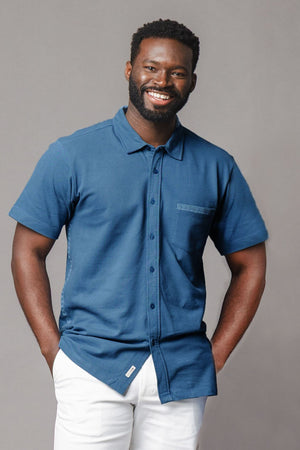 A smiling man models a short-sleeve stone blue shirt made of soft organic cotton, featuring a chest pocket and a relaxed fit, paired with light-colored pants.