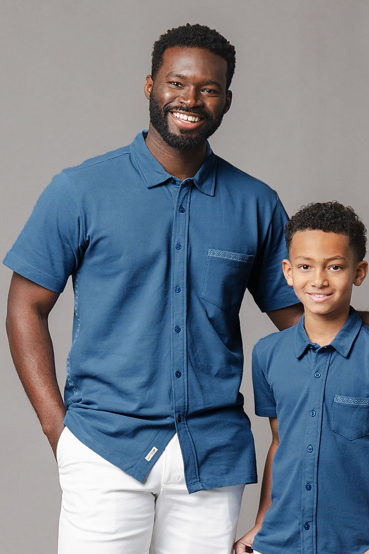 A smiling adult man and a boy wear matching stone blue short sleeve shirts with a pocket detail. The shirts are made from soft organic cotton, perfect for family outings.