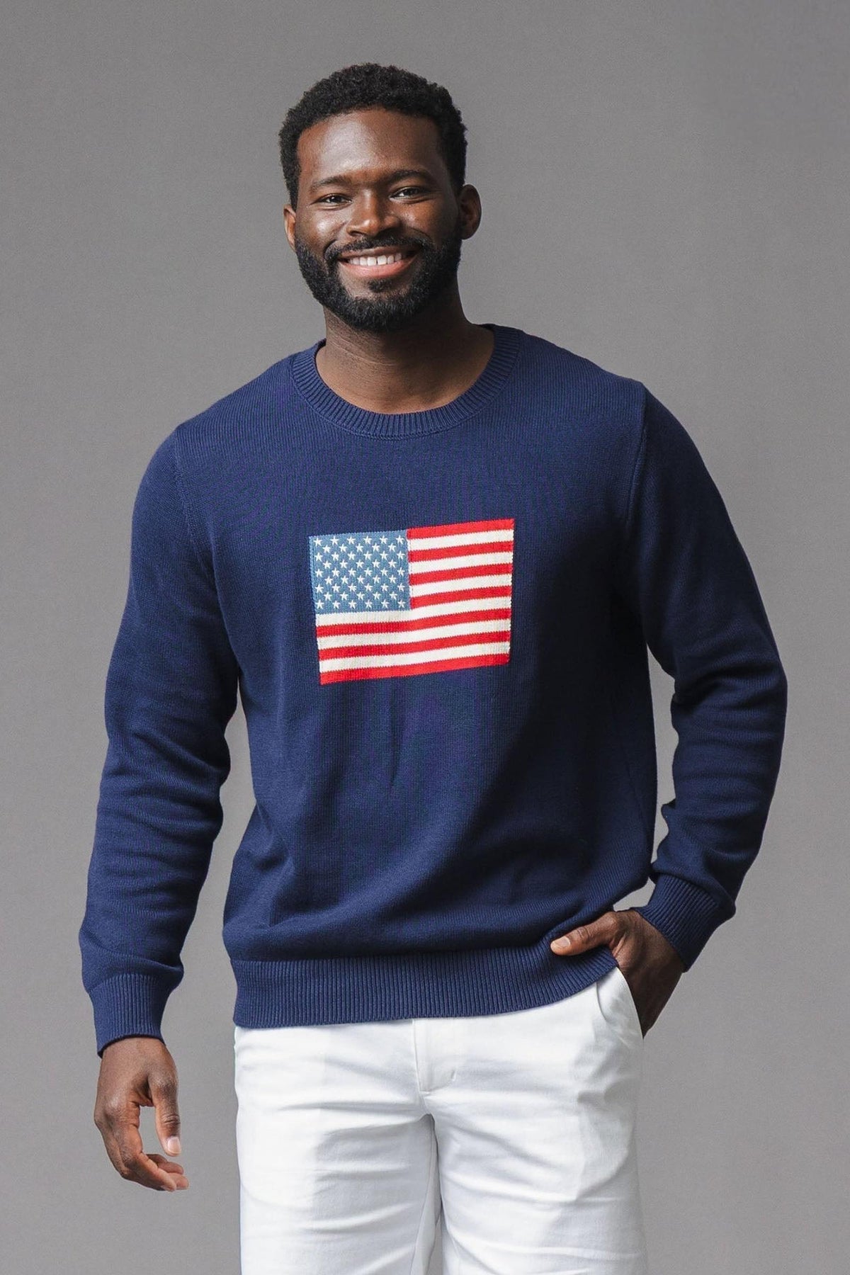 A man smiles while wearing a navy crew neck sweater featuring a prominent American flag design on the front. The sweater is made from soft, sustainable organic cotton.