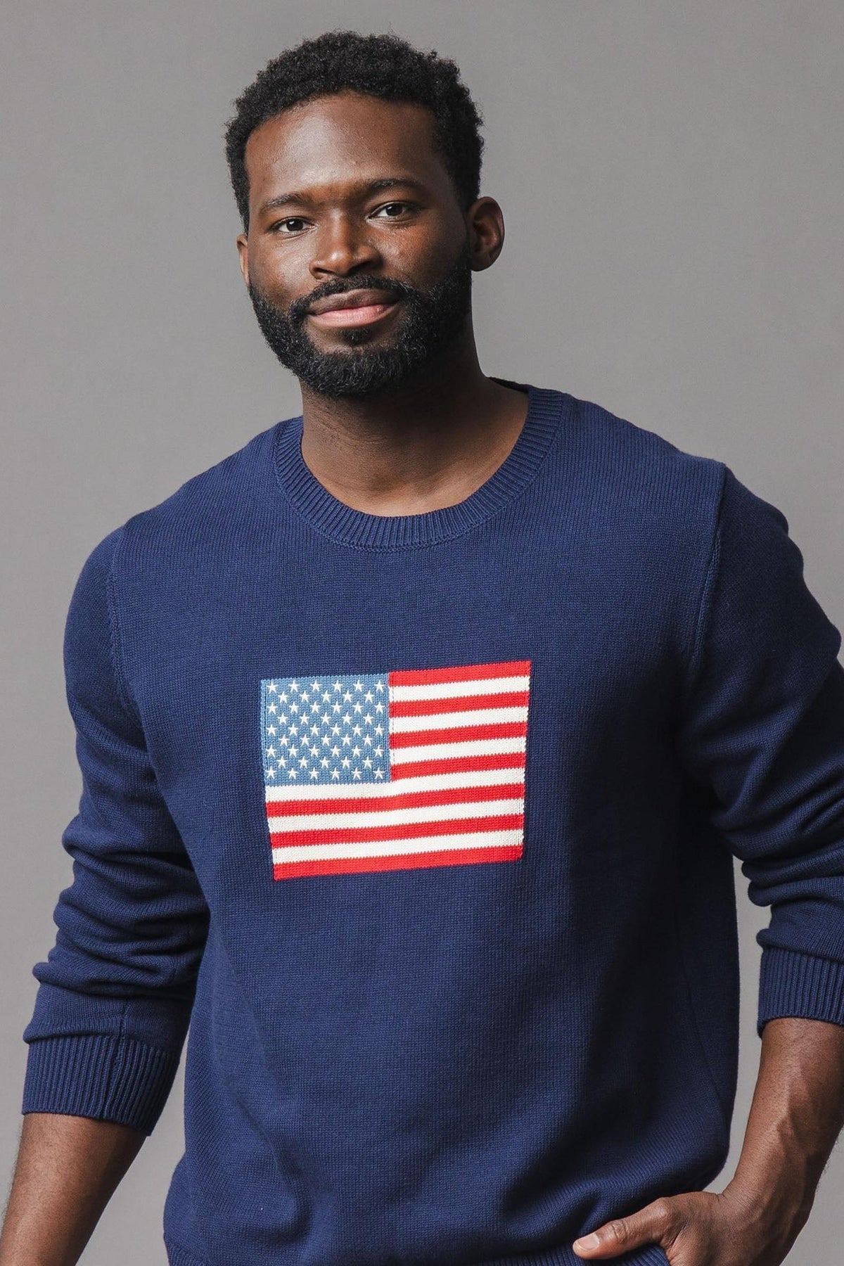 A man is wearing a navy blue sweater featuring a colorful American flag design on the front. The sweater has a classic crew neck style, made from soft, sustainable materials.