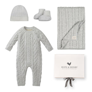 A light gray organic cotton cable romper, matching beanie, booties, and a blanket are neatly arranged beside a branded gift box. This sustainable baby gift set is perfect for newborns.