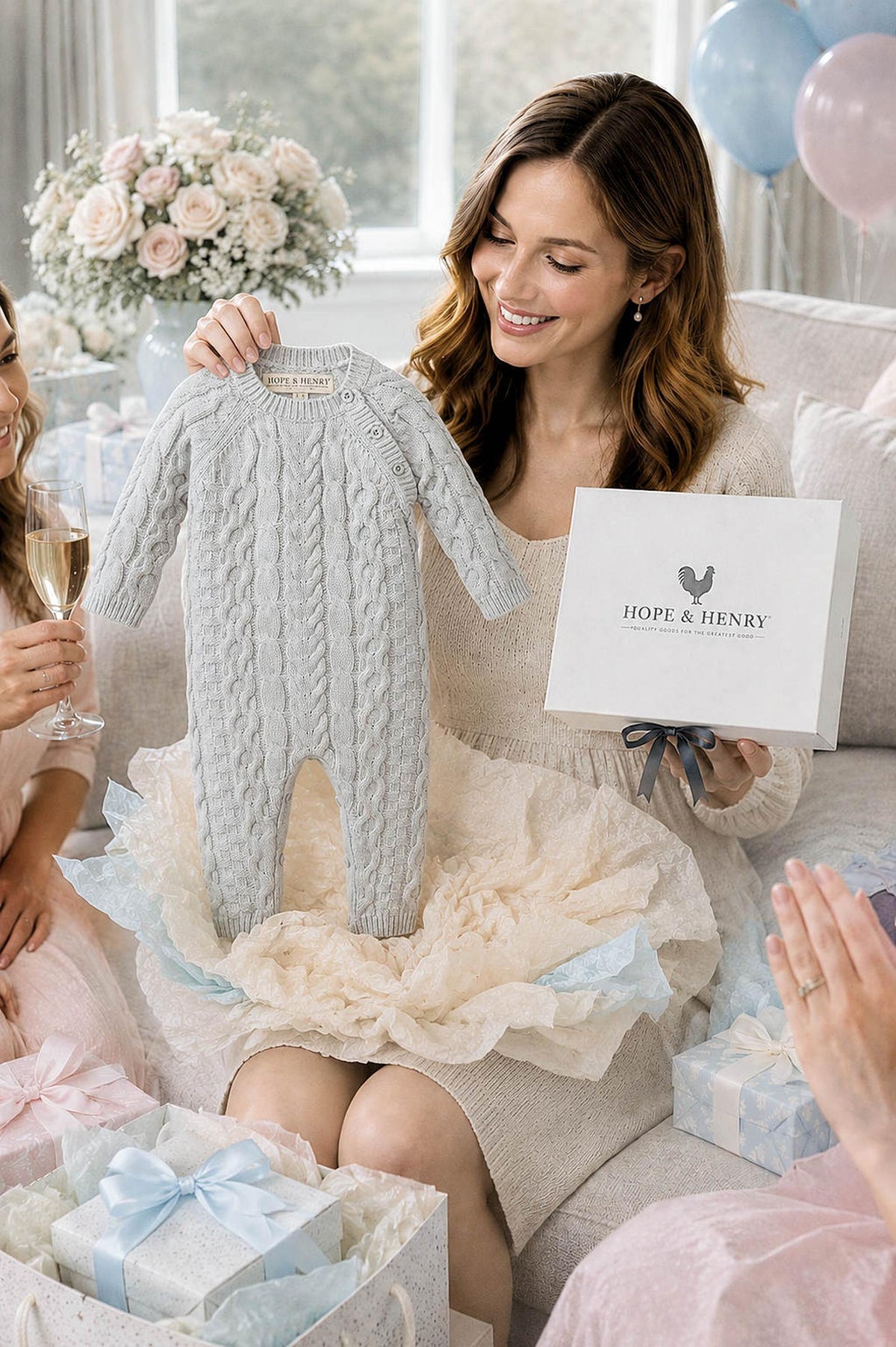 A woman holds a light gray cable-knit romper while sitting among friends at a gift-giving celebration. The romper is part of a beautifully packaged gift set from Hope & Henry.