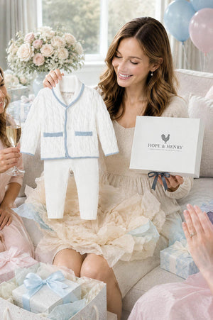 A woman in a light-colored dress smiles while holding a boxed gift set from Hope & Henry, featuring a white cable knit cardigan with blue accents and matching pants.