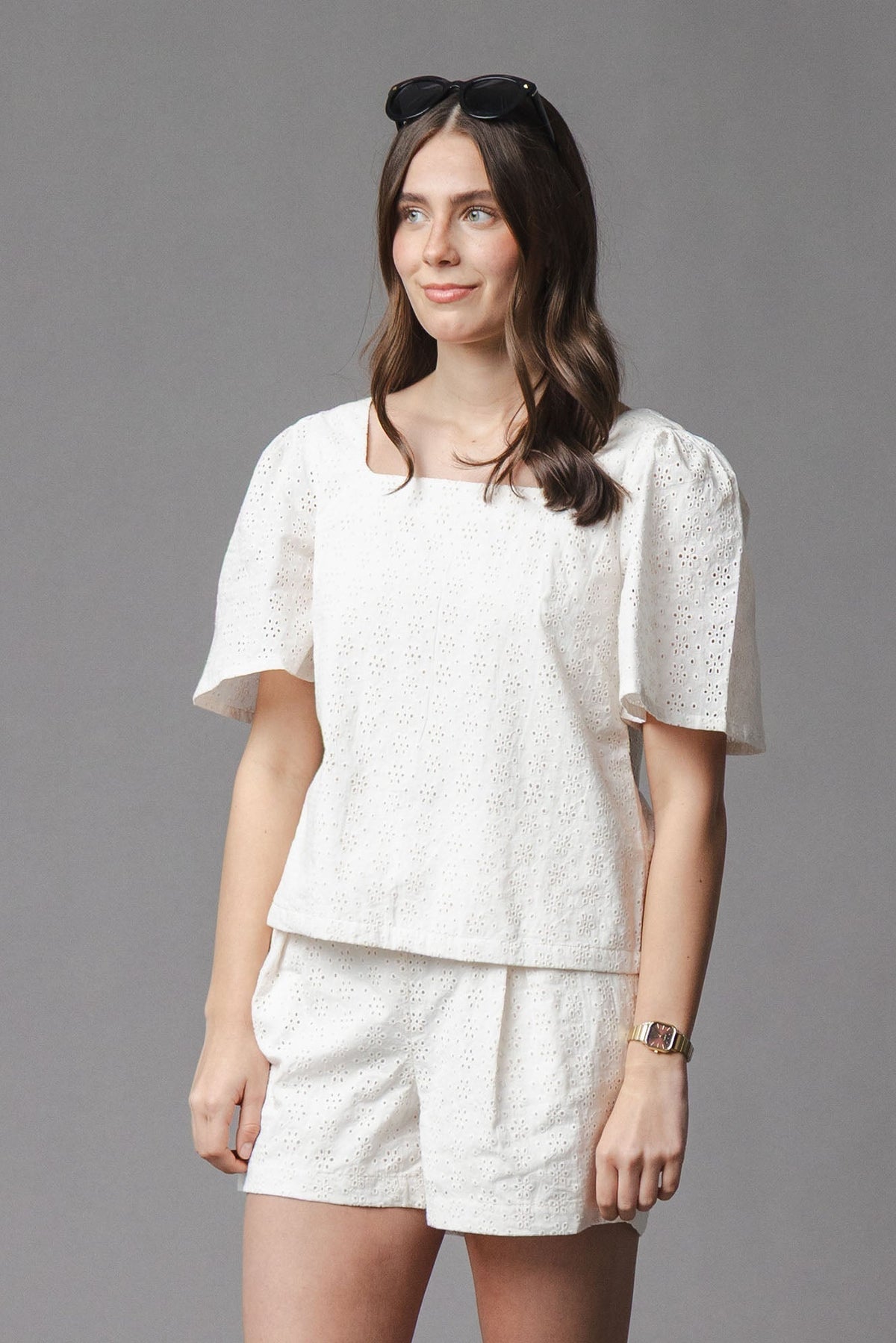 A woman wears a natural-colored organic cotton eyelet top with flounce sleeves, paired with matching shorts. The soft, breathable fabric is perfect for warm weather and family outings.