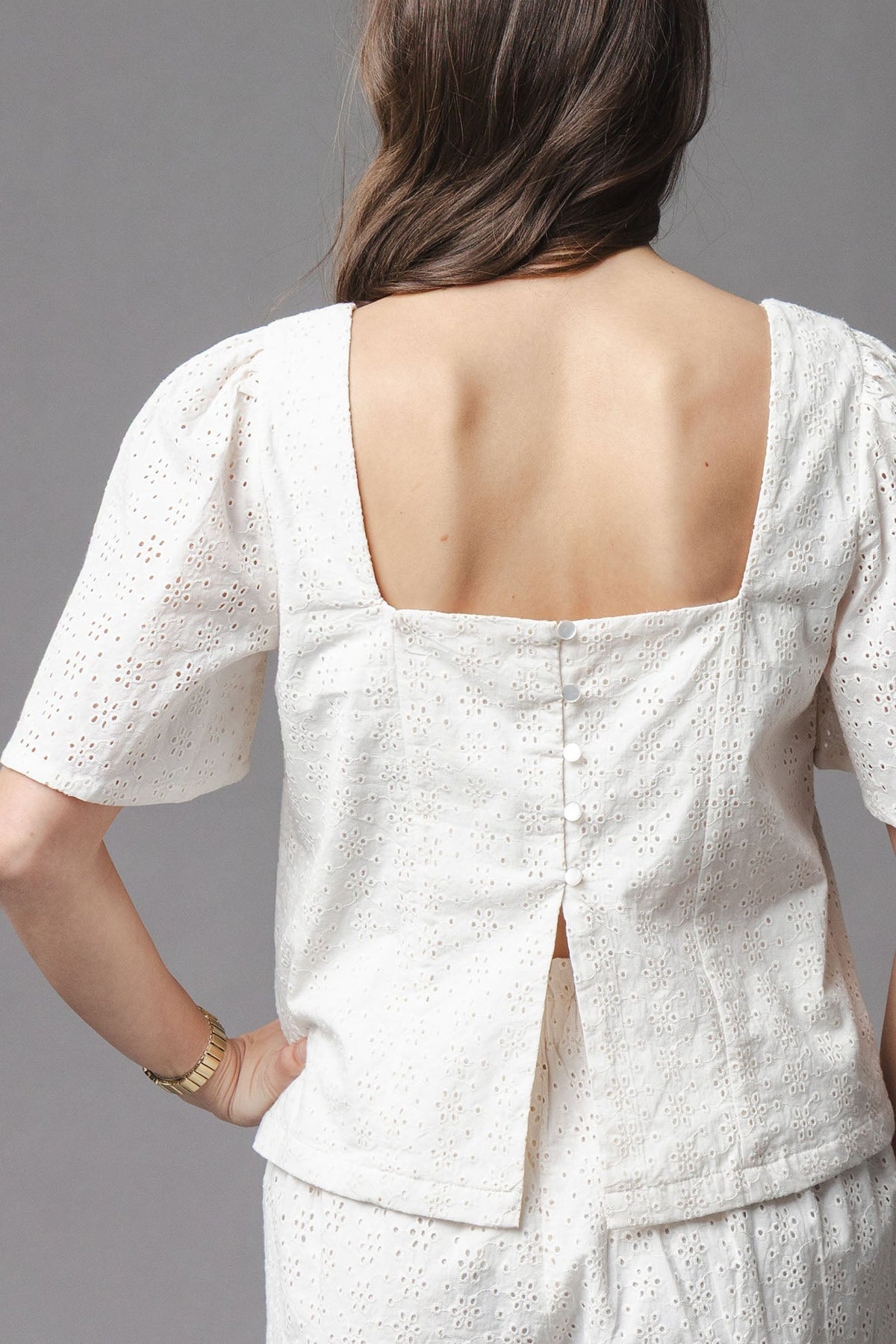 A woman is shown from behind wearing a natural-colored organic cotton eyelet top, featuring a square neckline and flounce sleeves, complemented by a button closure along the back.