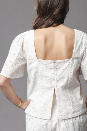 A woman is shown from behind wearing a natural-colored organic cotton eyelet top, featuring a square neckline and flounce sleeves, complemented by a button closure along the back.
