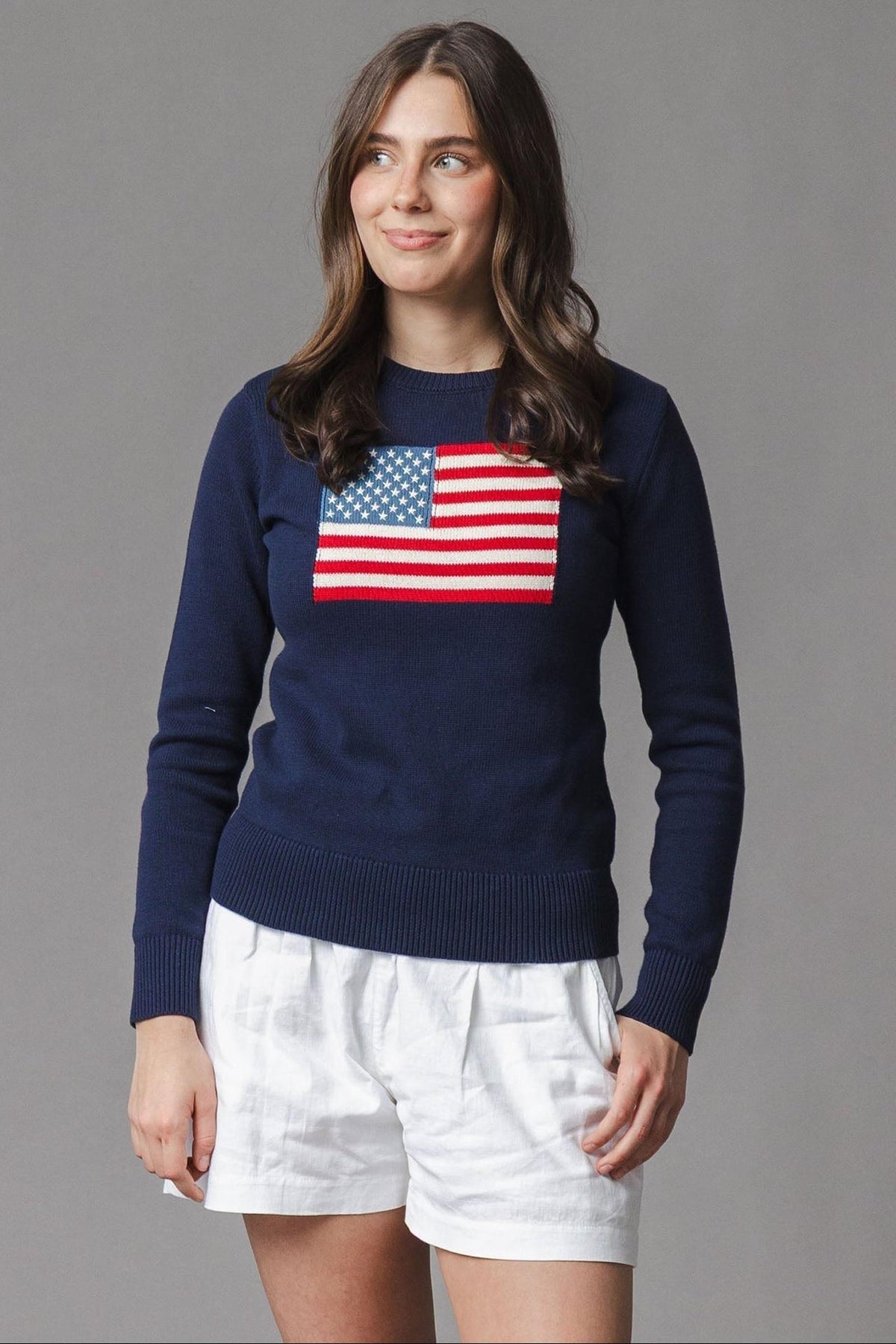 A woman stands wearing a navy organic cotton crew neck sweater featuring a classic American flag design. She pairs it with white shorts, set against a neutral gray backdrop.