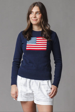A woman stands wearing a navy organic cotton crew neck sweater featuring a classic American flag design. She pairs it with white shorts, set against a neutral gray backdrop.
