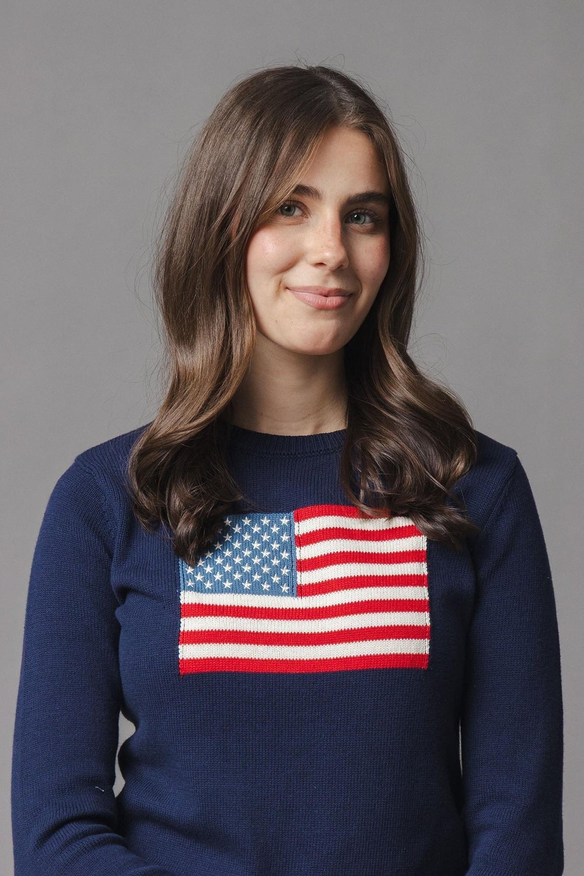 A woman with long, wavy hair wears a navy sweater featuring a classic American flag design on the chest. The sweater is crafted from soft, organic cotton for comfort.