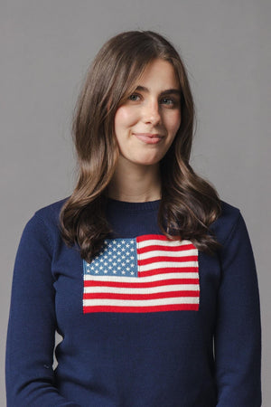 A woman with long, wavy hair wears a navy sweater featuring a classic American flag design on the chest. The sweater is crafted from soft, organic cotton for comfort.