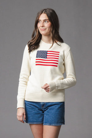 A woman models an ivory organic cotton crew neck sweater featuring an American flag design. The sweater is paired with denim shorts, set against a neutral gray background.