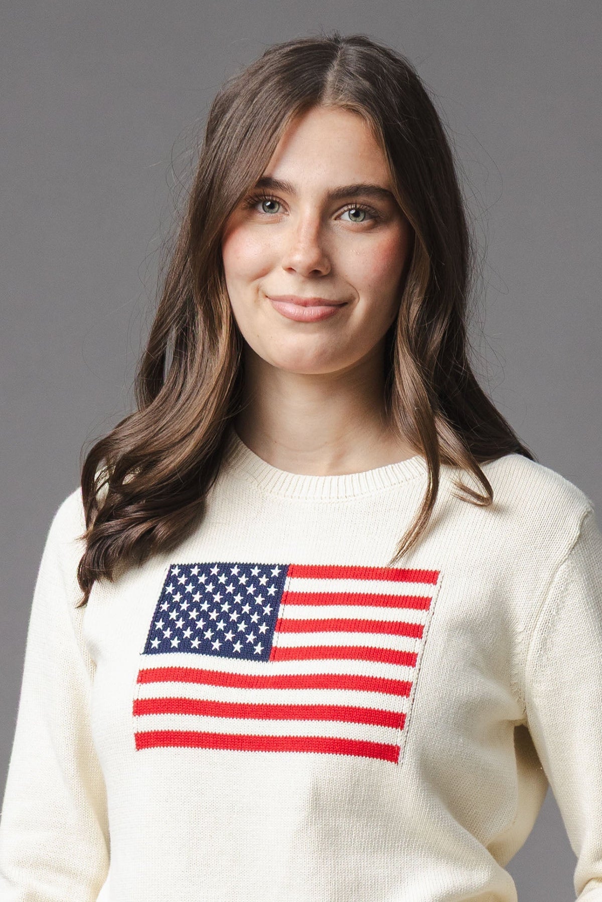 A young woman smiles while wearing an ivory crew neck sweater featuring a red and blue American flag design on the front. The sweater is made from soft organic cotton.