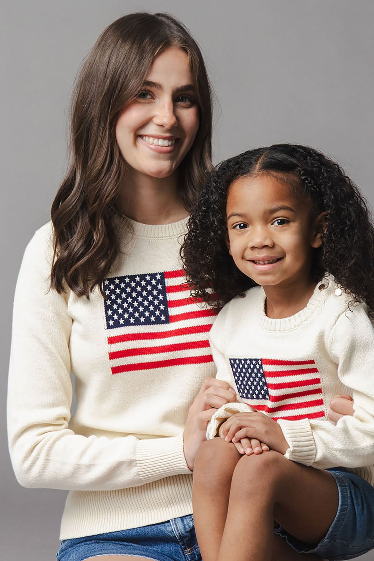 A woman and a young girl smile while wearing matching ivory crew neck sweaters featuring an American flag design.