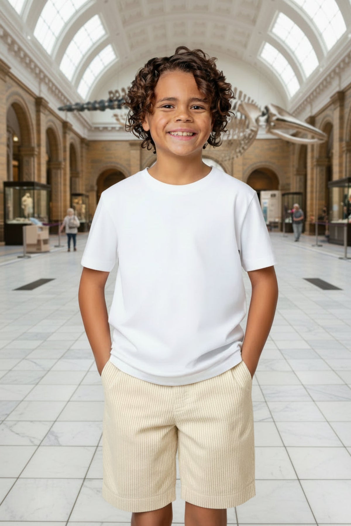 A smiling boy stands in a museum atrium, wearing a white t-shirt and light-colored seersucker shorts. The spacious background features exhibits and large windows, creating a bright atmosphere.
