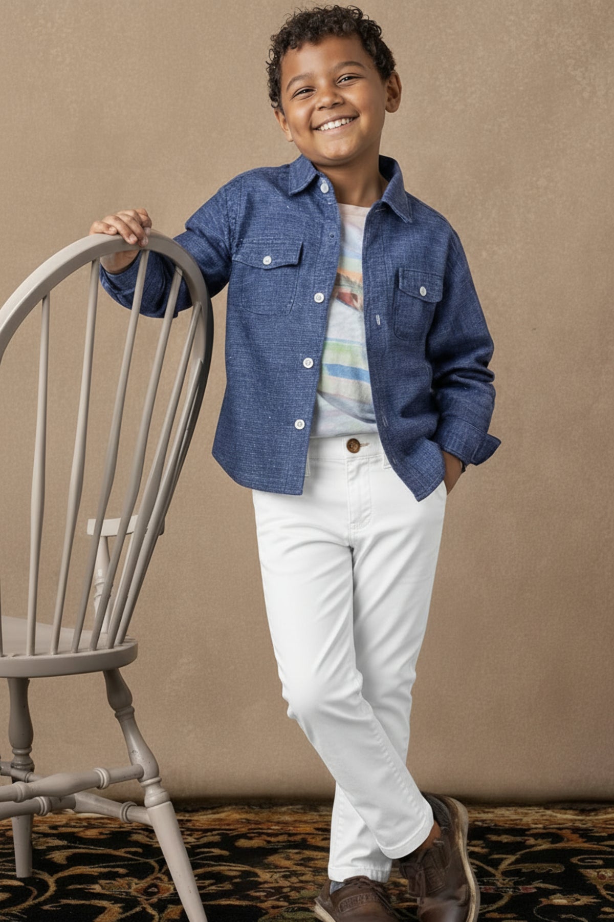 A young boy wearing a blue shirt and white chinos smiles while leaning on a wooden chair.