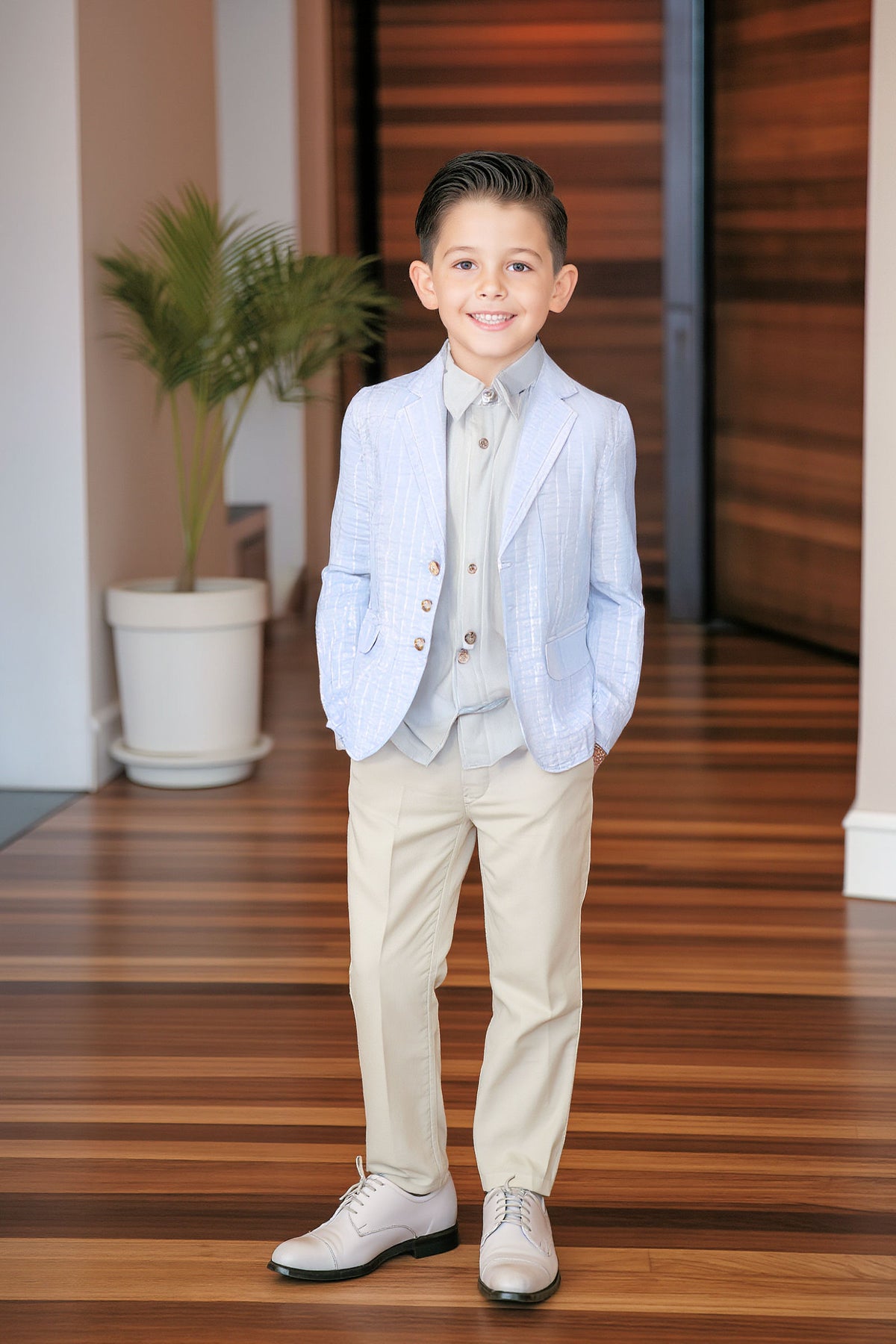 A smiling boy wears a light blue seersucker blazer, beige pants, and white shoes in a modern hallway.