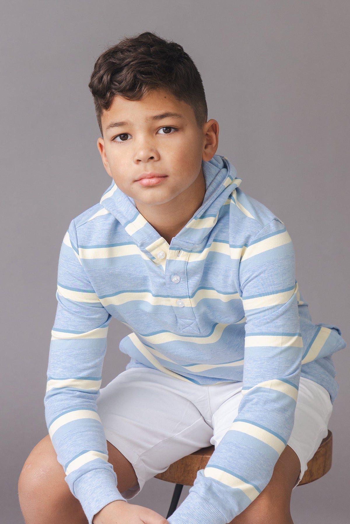 A young boy sits on a wooden stool, wearing a blue and yellow striped organic cotton hoodie.